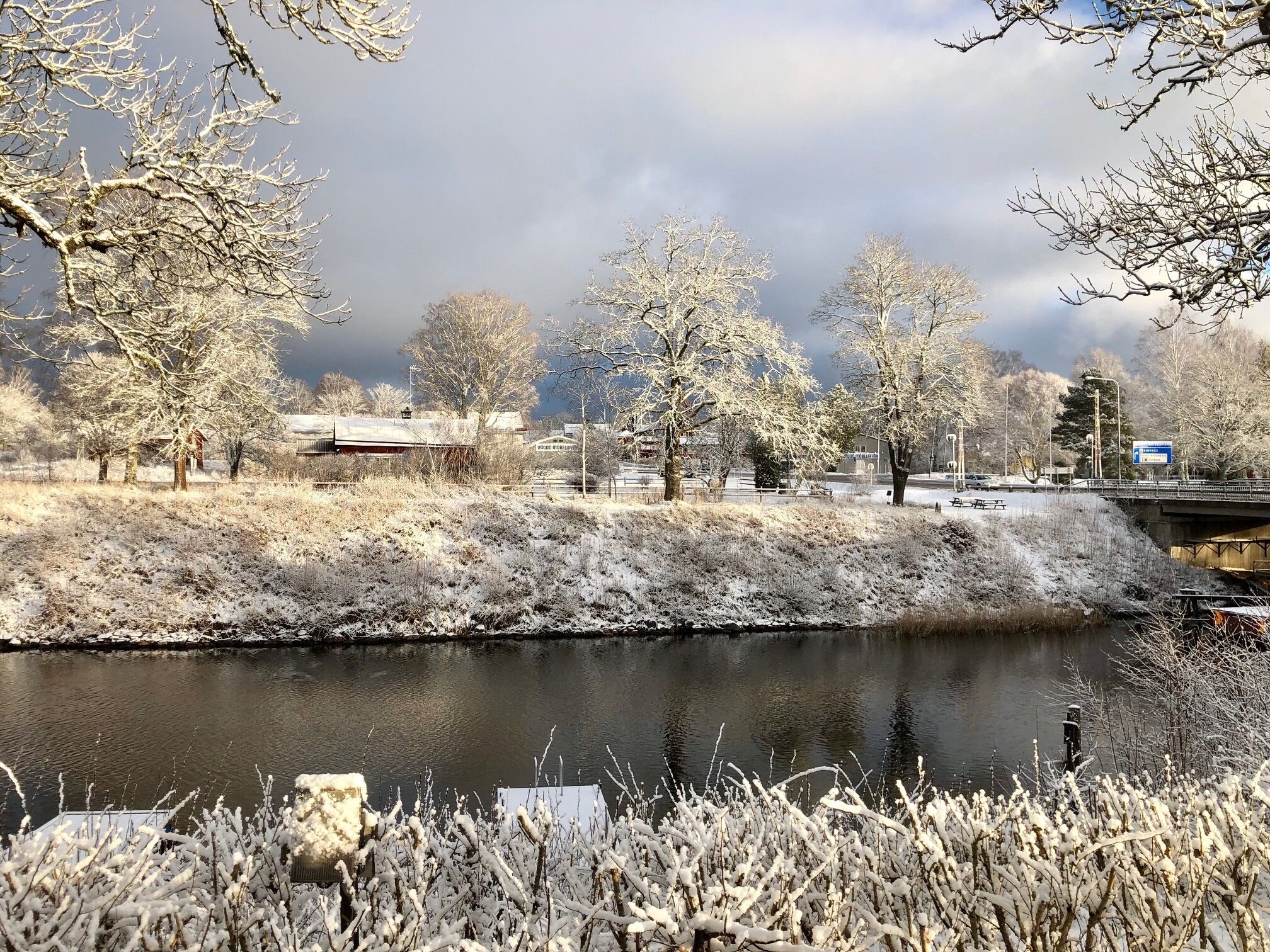 Een winterlandschap met een bevroren rivier, kale bomen en een brug op de achtergrond.