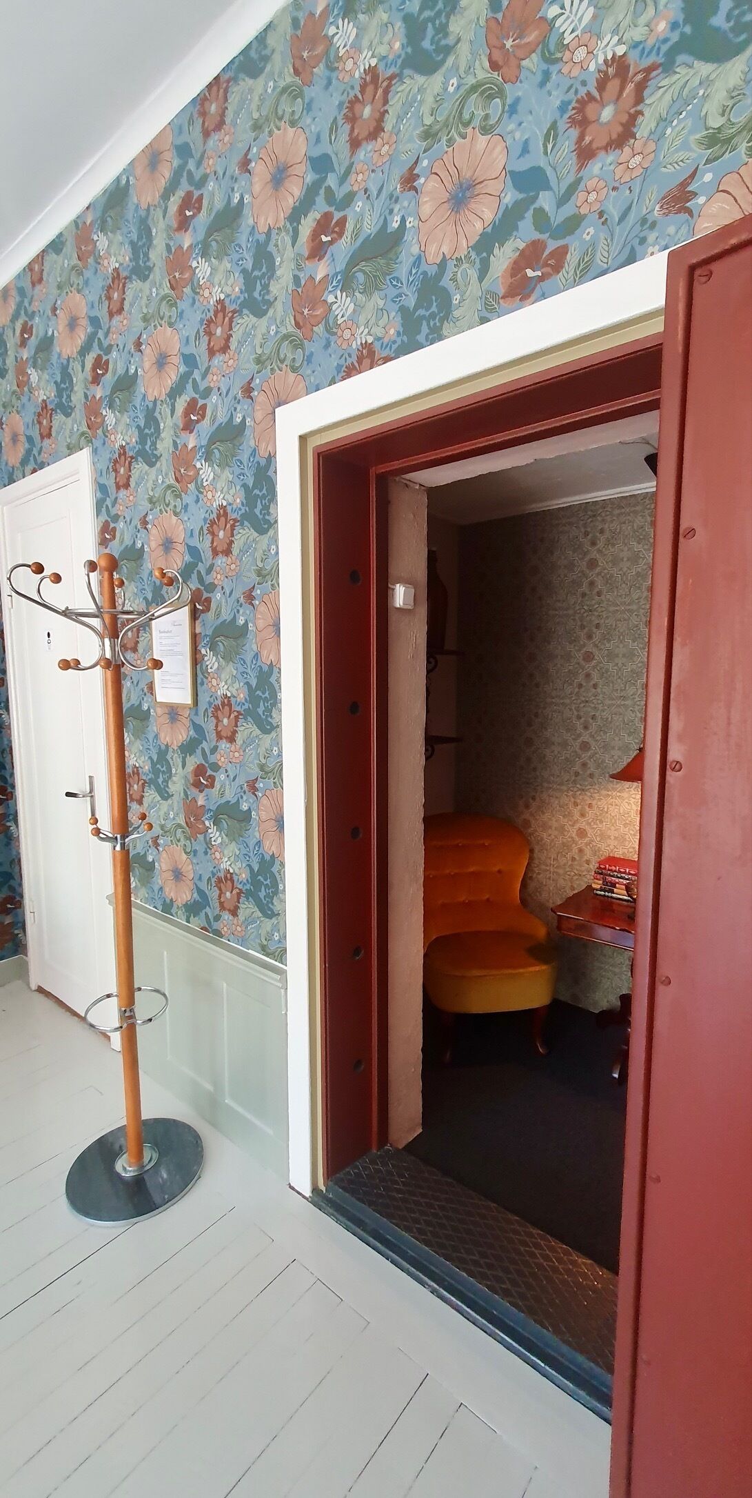 A room with a floral wallpaper, a wooden coat rack, and a red door leading to another room.