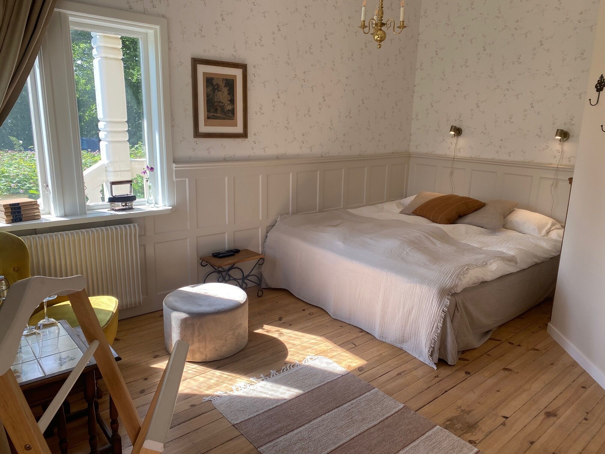 A bedroom with a bed, a small table, a chair, a radiator, and a window with curtains.