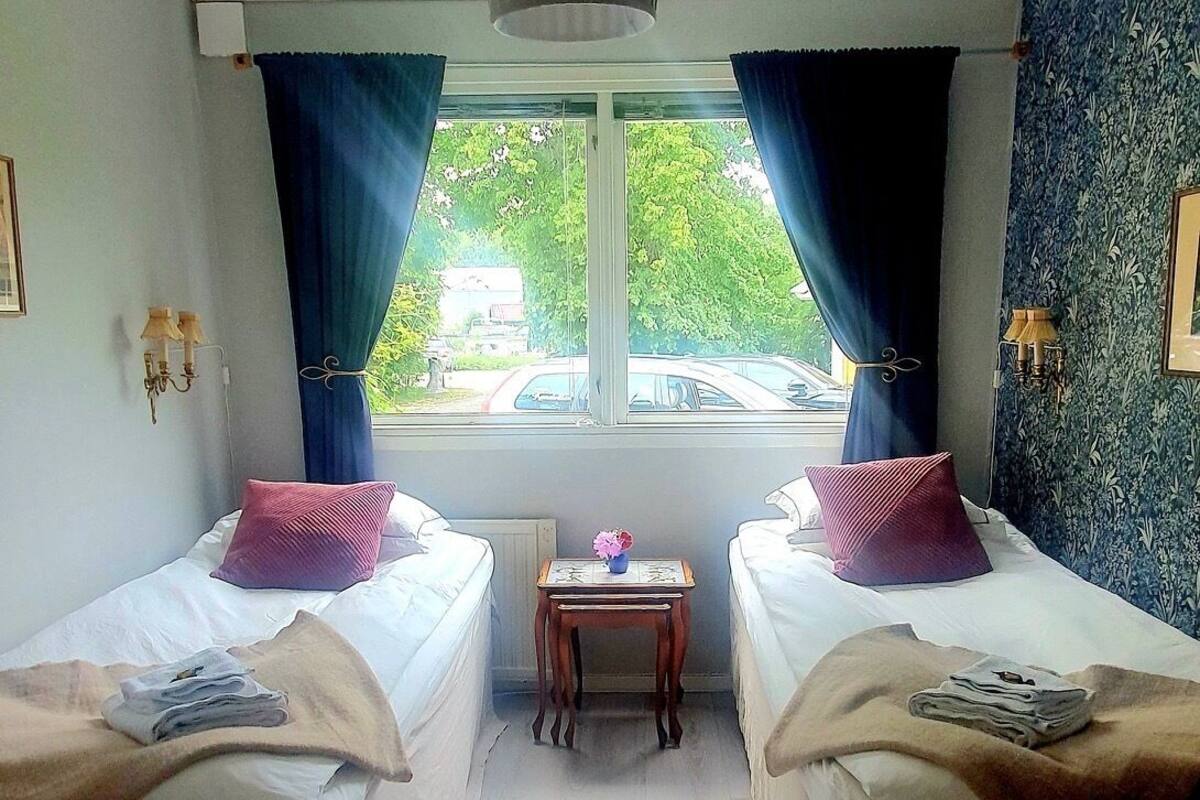 A room with two single beds, a small table, a hanging light fixture, and a window with curtains.