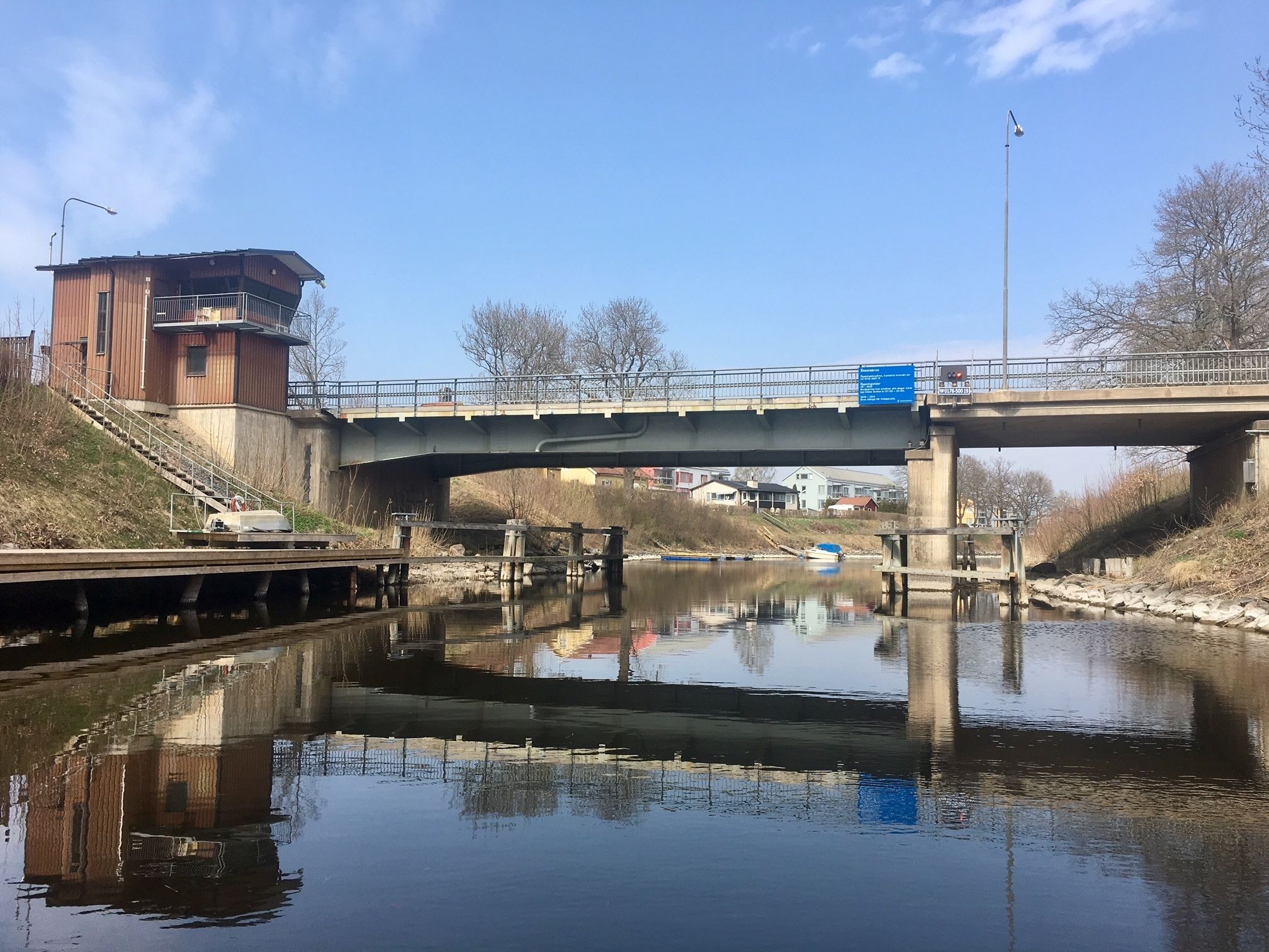 A bridge over a calm river with a reflection of the bridge and buildings. There is a small building on the left and a staircase leading up to it.
