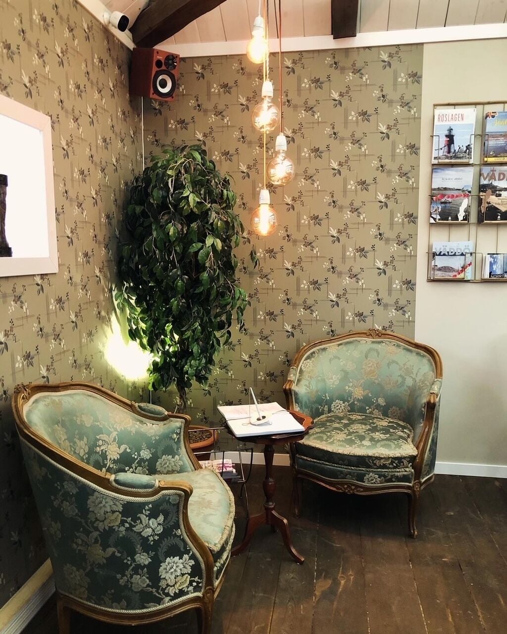 A room with floral wallpaper, two vintage-style chairs, a small table, and a large green plant.