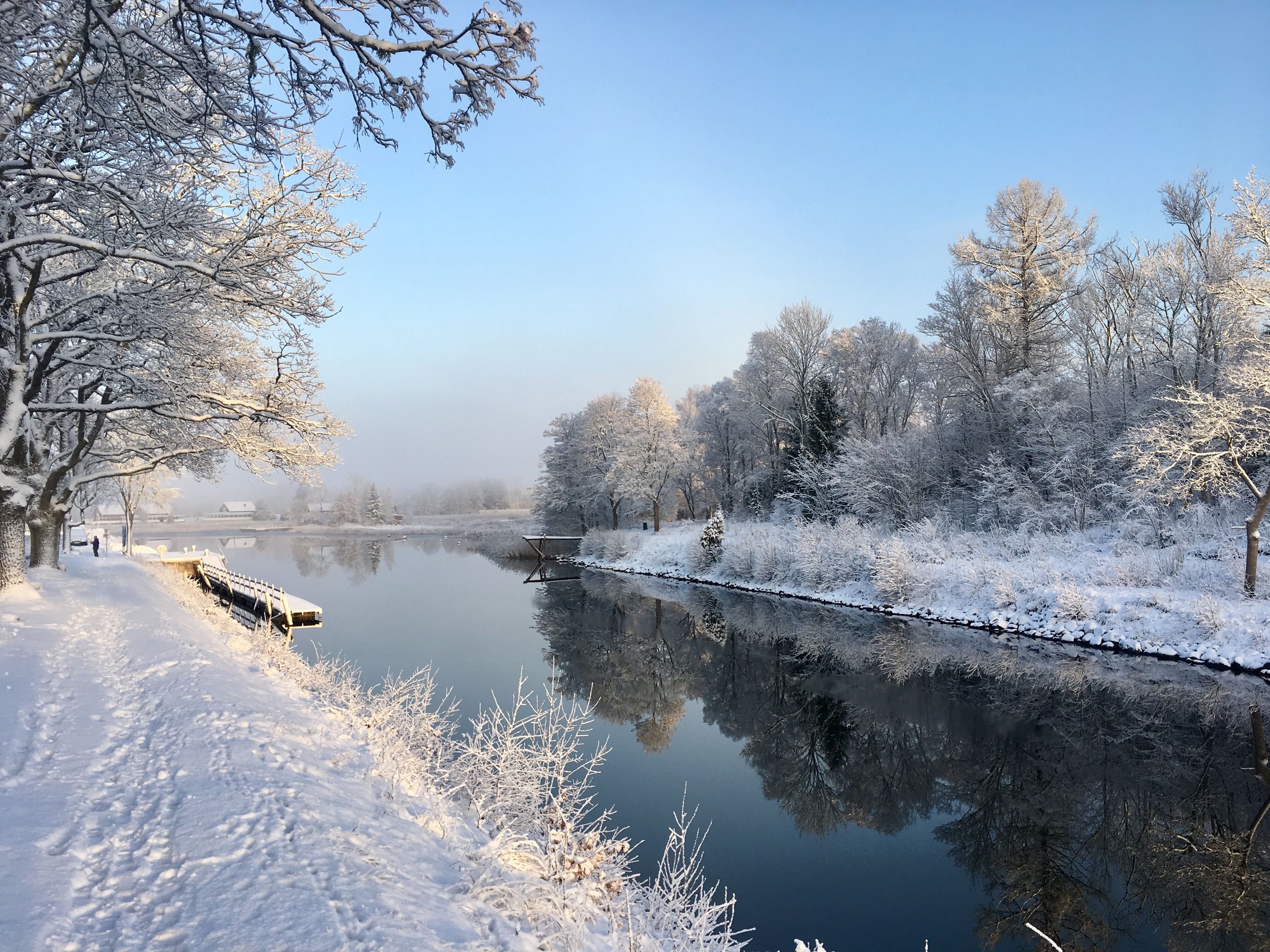 A serene winter scene with a snow-covered river, trees, and a wooden bridge.