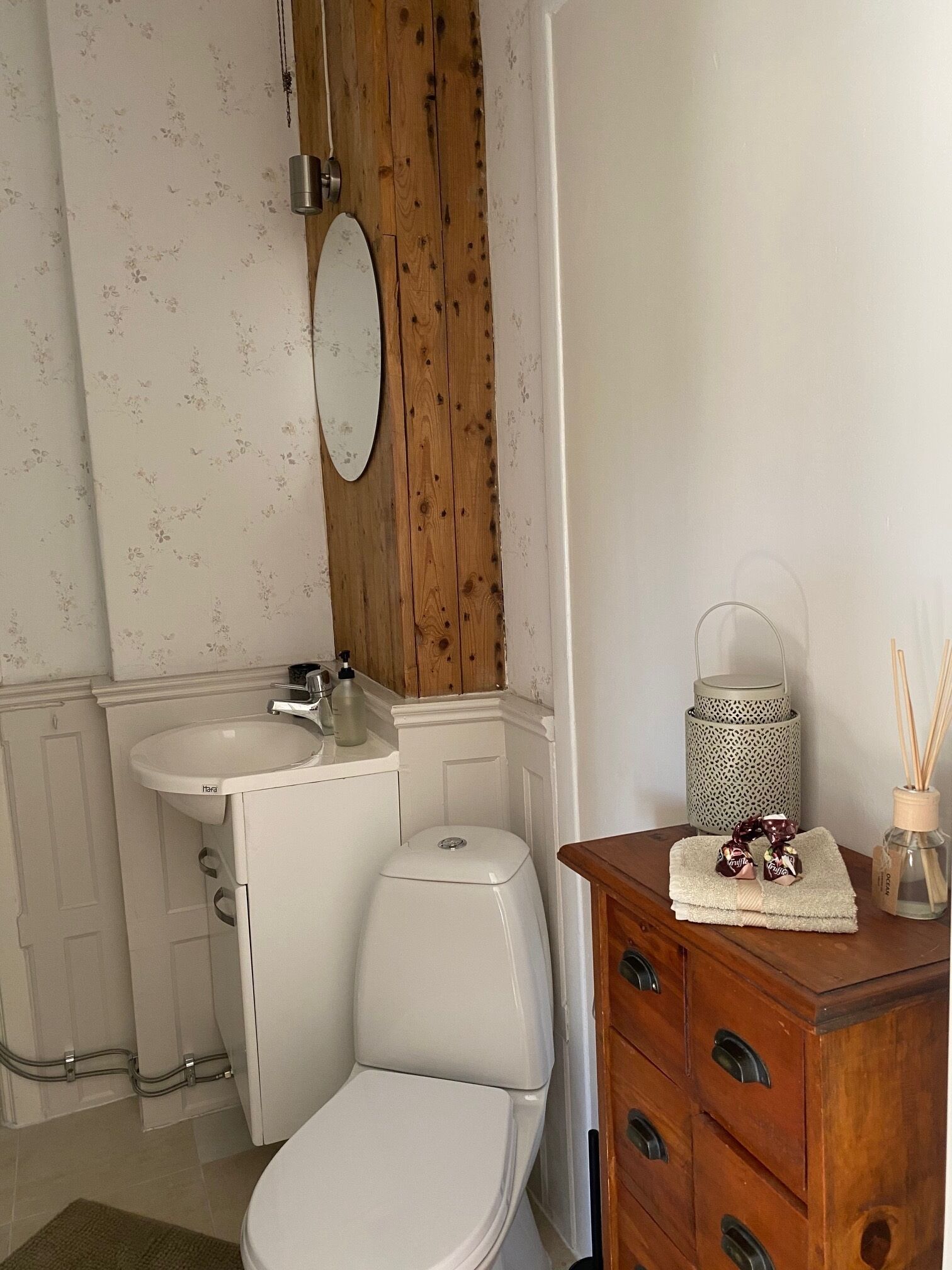 Un baño con inodoro, lavamanos y espejo. Se observa un mueble de madera con cajones y una mesita con una botella y una vela.
