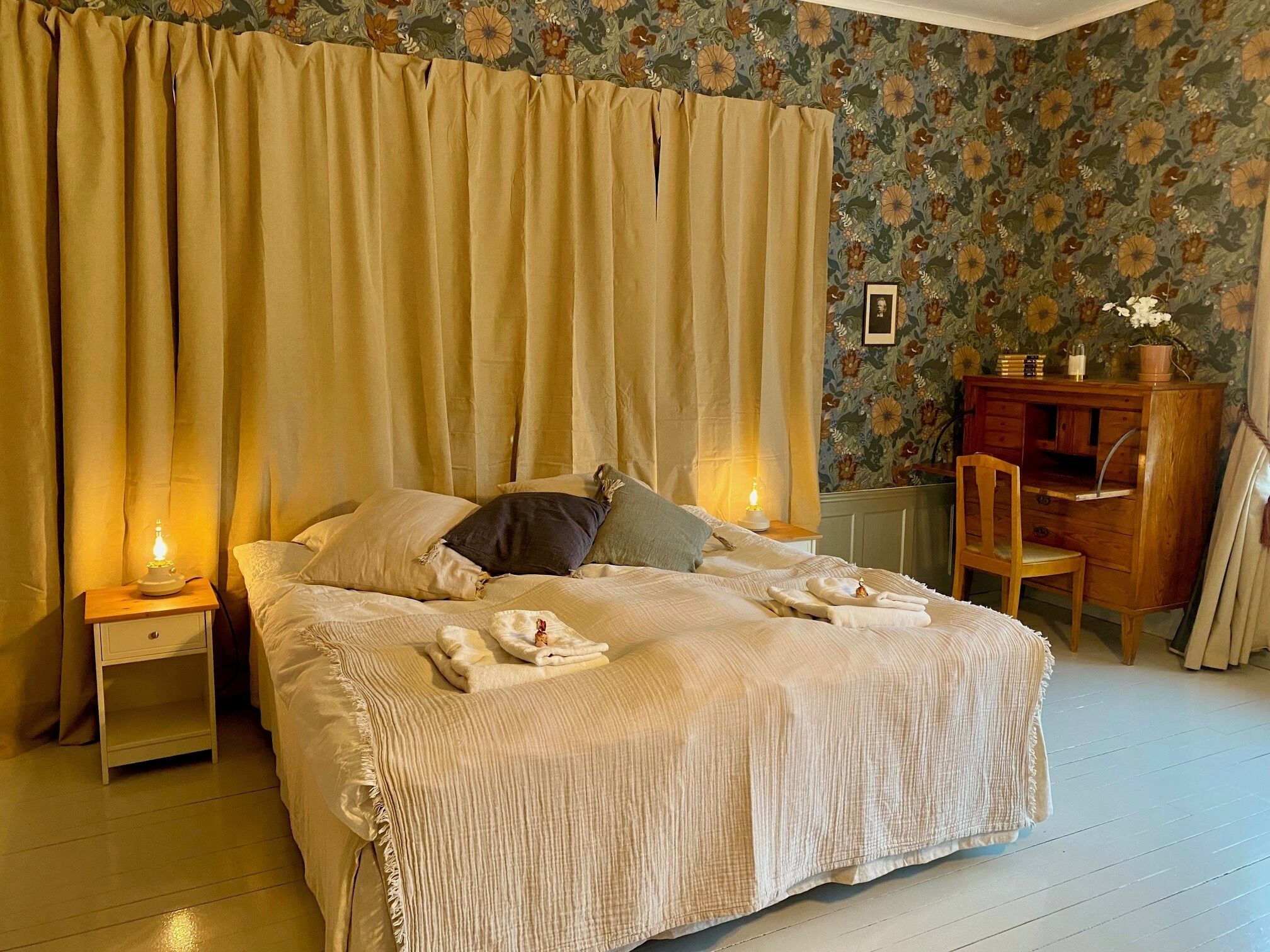 Una cama bien tendida con cubrecama y almohadas color beige, dos lámparas de noche, un escritorio de madera con una silla, y un estampado floral en el papel tapiz.