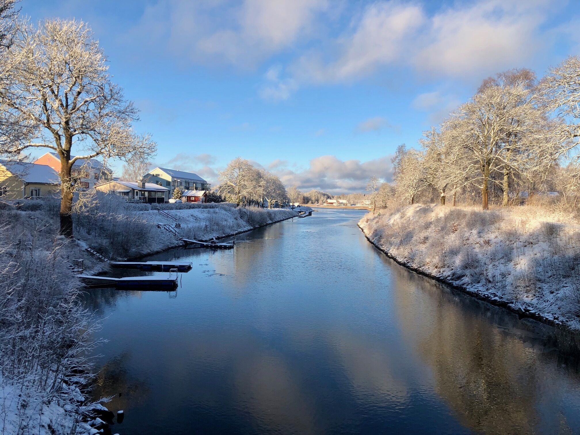 A river with snow-covered banks and trees, houses along the riverbank, and a clear sky.