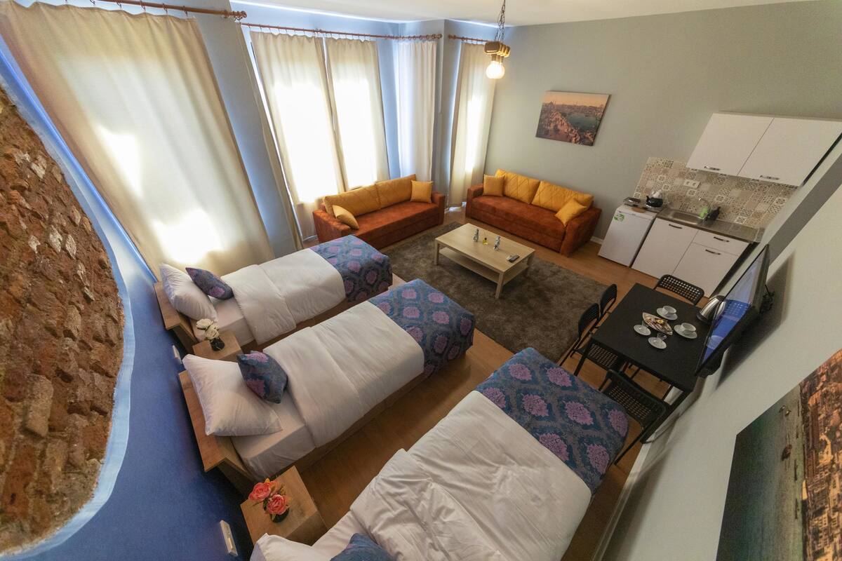 A compact hotel room with two beds, a sofa, a small table, and a kitchenette.