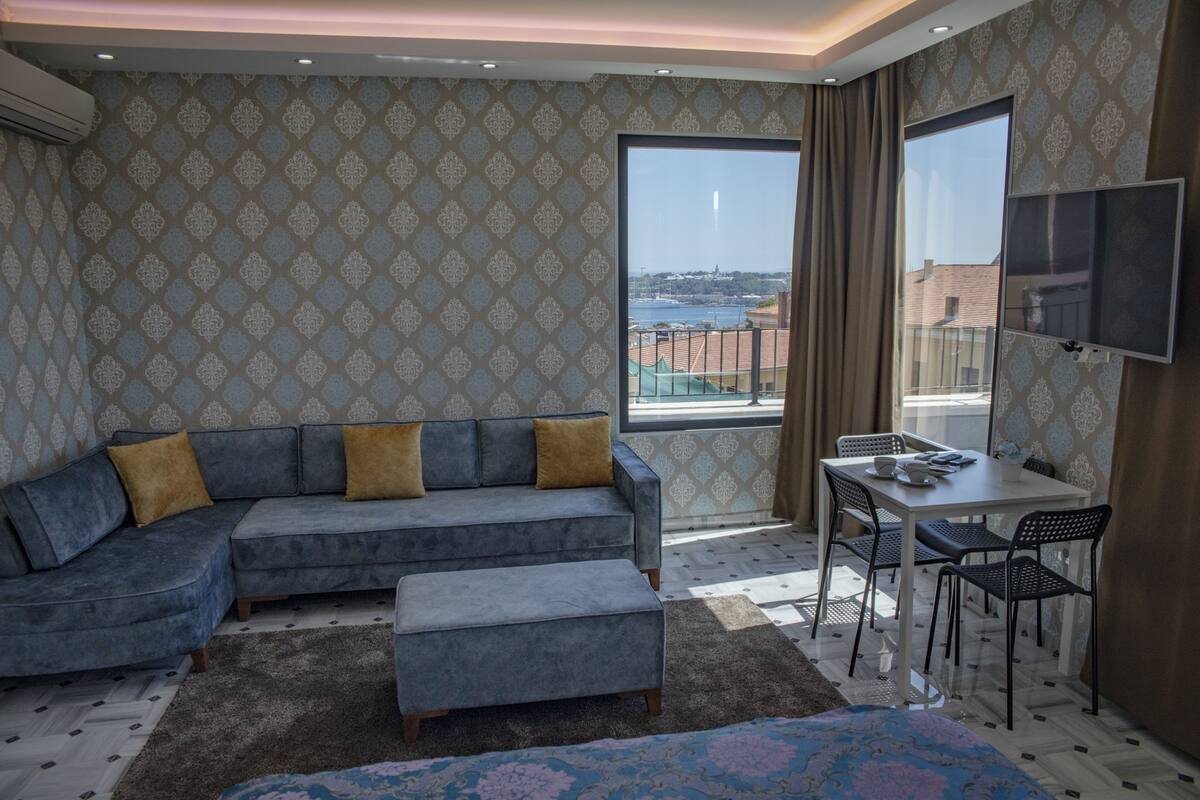 A modern living room with a grey sectional sofa, a small table with chairs, and a large window offering a view of a cityscape.