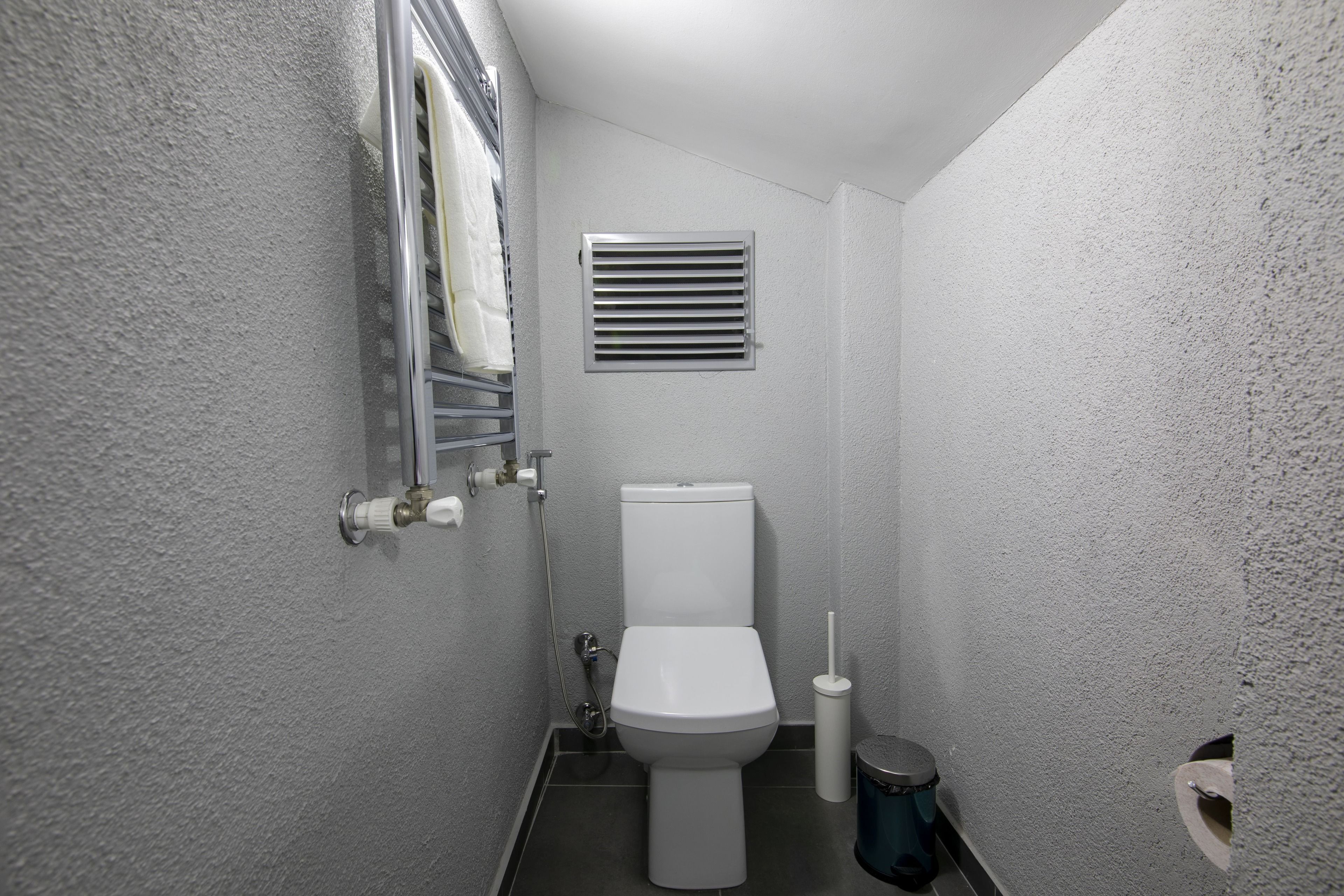 A small, compact bathroom with a white toilet, a wall-mounted vent, and a towel rack.