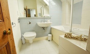 A bathroom with a toilet, sink, and bathtub. There is a wooden door with a brass handle, and a small trash can is visible.