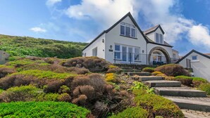 A two-story house with a stone staircase leading to the entrance, surrounded by lush greenery and a hill in the background. - Stables Croyde 4 Bedrooms, Sleeps 8, Sea & Beach Views (Braunton)