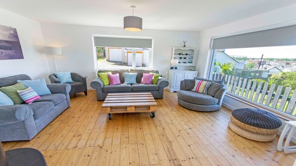 A modern living room with grey sofas, a wooden coffee table, and a view of a garden through large windows.