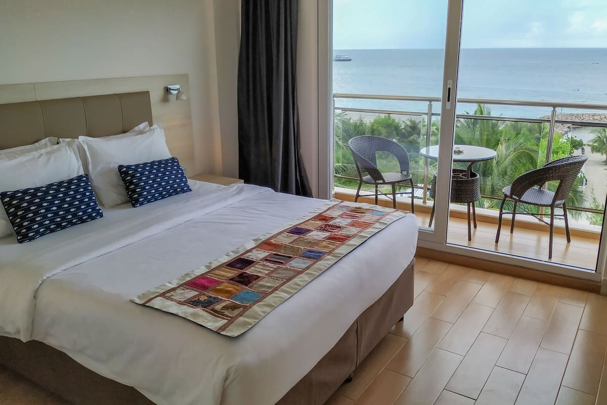 A hotel room with a large bed, a view of the sea, and a balcony with chairs.