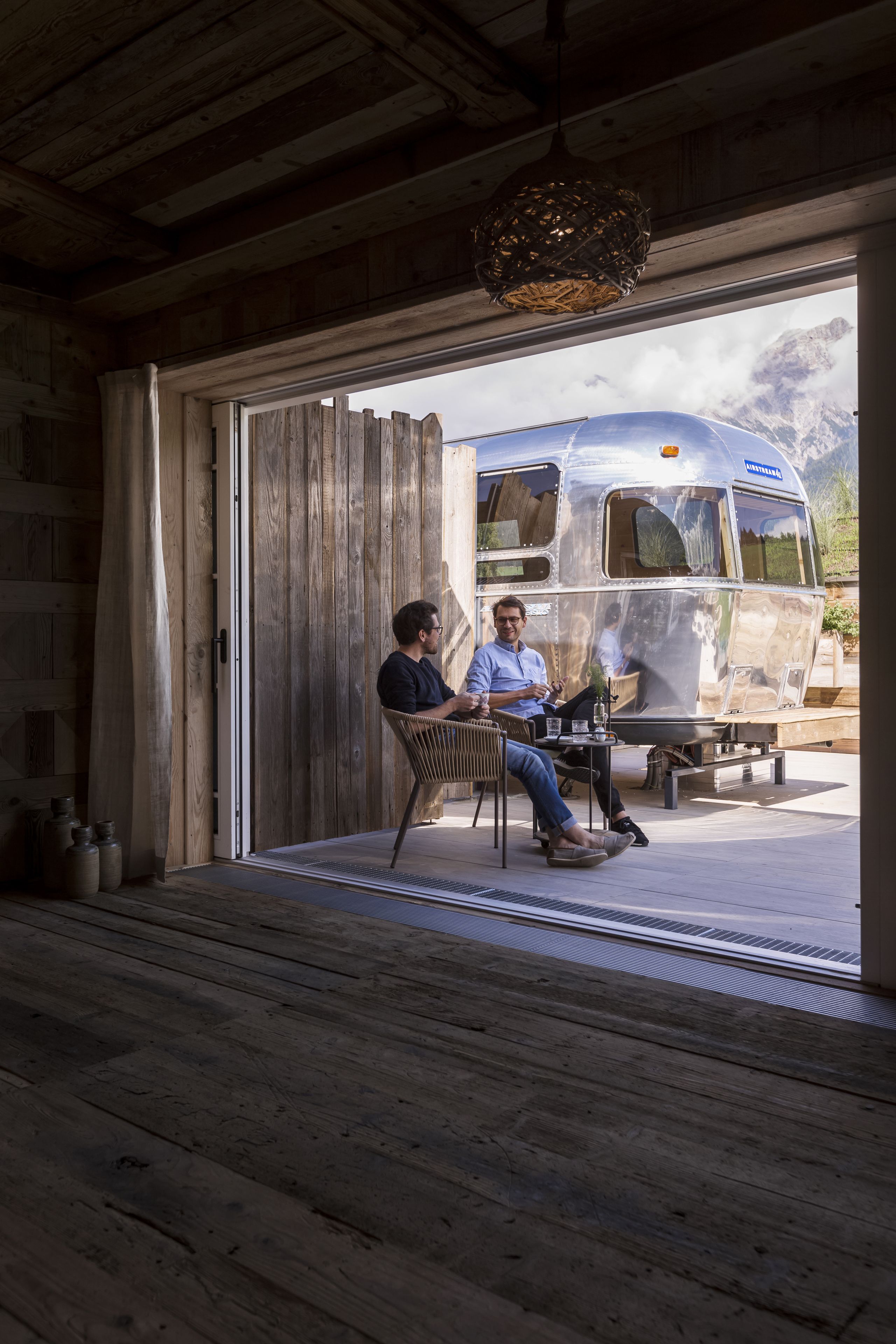Un interior rústico con techo de madera y un amplio ventanal que muestra una caravana antigua estacionada afuera.