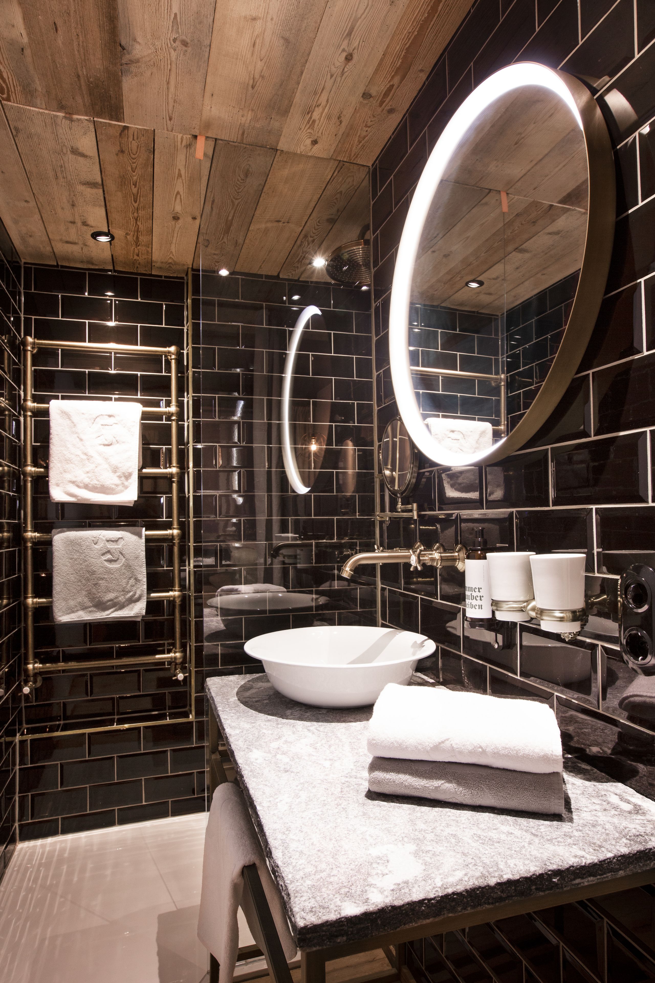 A modern bathroom with a wooden ceiling, black tiled walls, a large mirror, a white sink, and a towel rack.