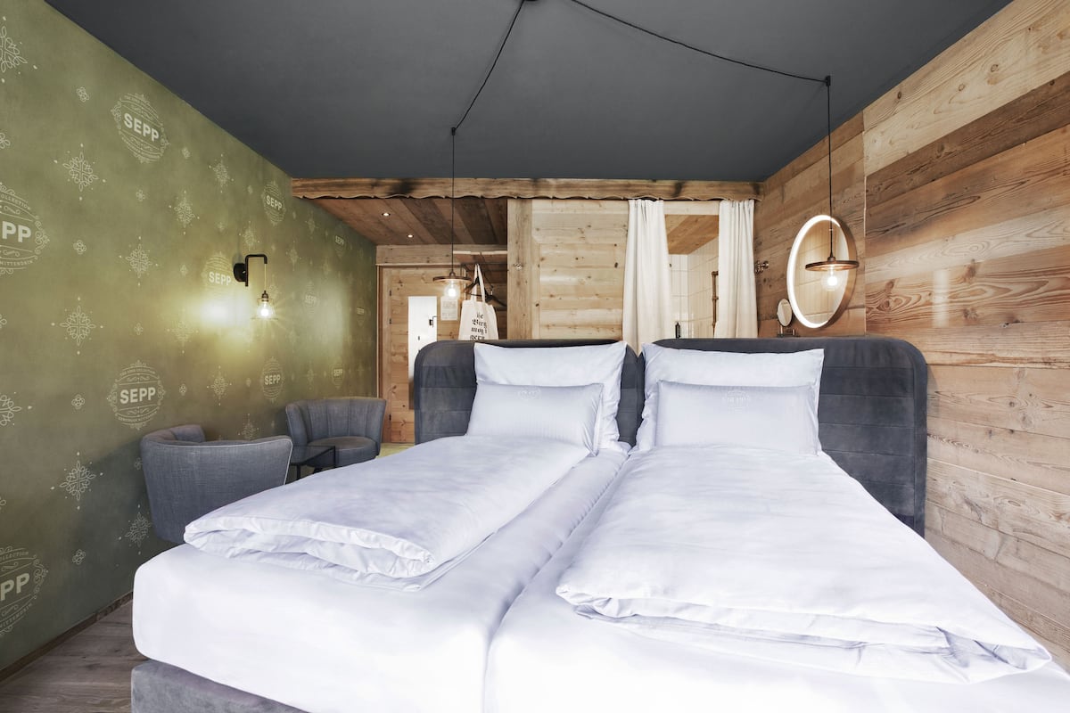 Ein Zimmer mit zwei Betten, einer Holzwand, einer grünen Wand mit SEPP-Branding, einer Pendelleuchte und einem Spiegel.