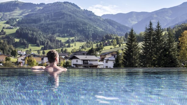 A person in a swimming pool with a view of houses and mountains in the background. - SEPP - Alpine Boutique Hotel (Maria Alm am Steinernen Meer)