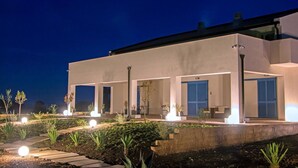 A modern building with a flat roof, large windows, and a well-lit outdoor area with plants and a pathway. - Melus Maris (Syracuse)