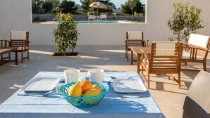 An outdoor dining area with a table set for two, a fruit basket, and a view of a swimming pool and garden. - Melus Maris (Syracuse)