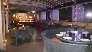 An outdoor dining area with wicker furniture and tables set for two, featuring a brick wall and a bar area in the background. - Kyriad Hotel Indore (Indore)