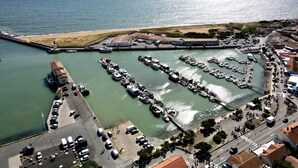 Vista aérea de uma marina costeira com barcos atracados, um farol e uma estrada que leva a uma cidade.