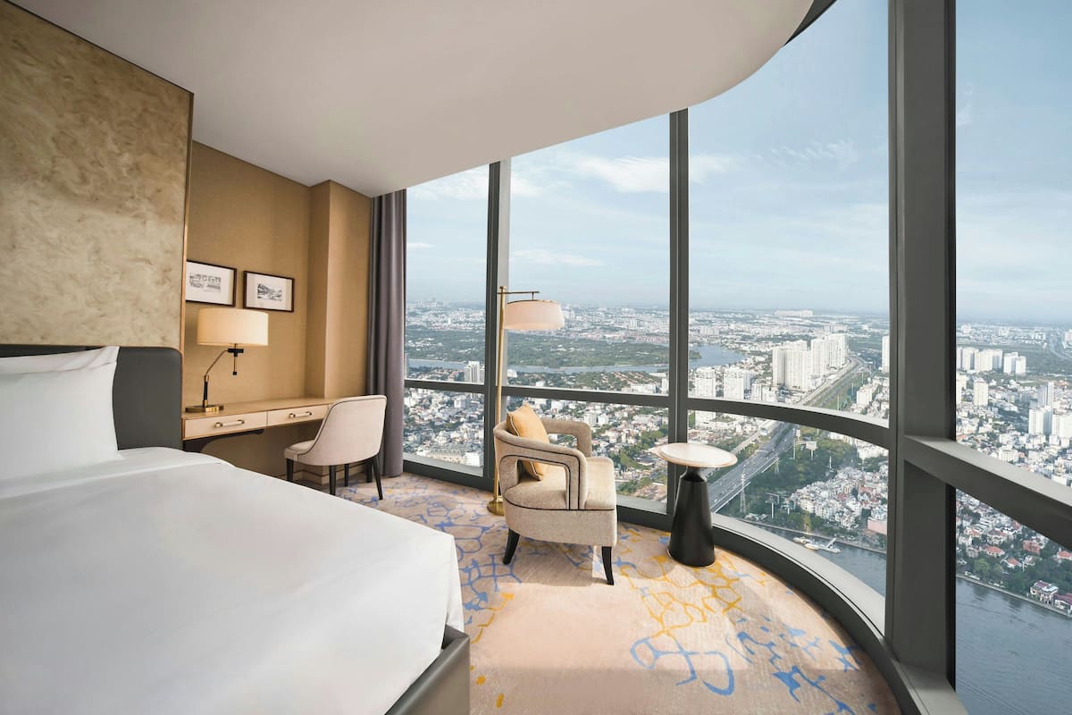Habitación de hotel con una cama grande, un escritorio, una silla y vistas de la ciudad a través de amplias ventanas.