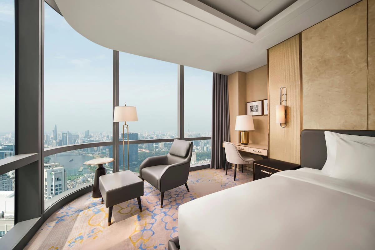 Habitación de hotel con una cama grande, un escritorio, una silla y vistas a la ciudad a través de amplios ventanales.