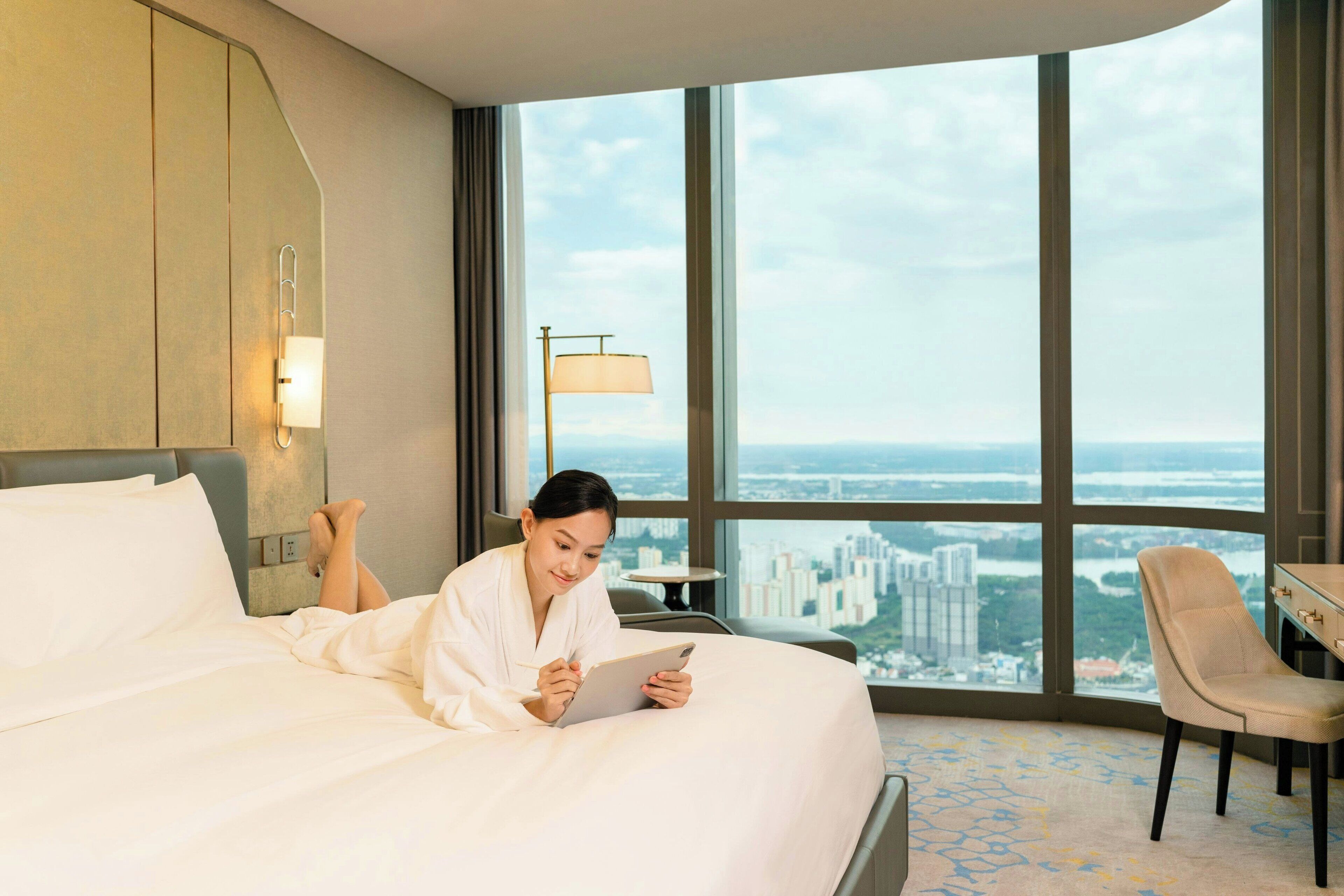 Une chambre d’hôtel avec un grand lit, une vue sur le paysage urbain, un bureau avec une lampe et une chaise.