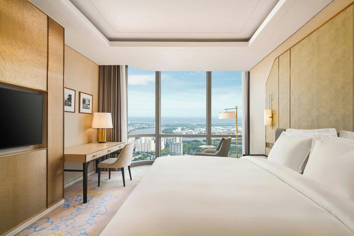 Habitación de hotel con una cama grande, un escritorio con silla, televisión y vistas a un paisaje urbano a través de ventanas amplias.