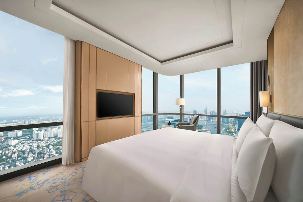 Habitación de hotel con una cama grande, un televisor de pantalla plana en la pared, una silla y vistas a un paisaje urbano a través de ventanas amplias.