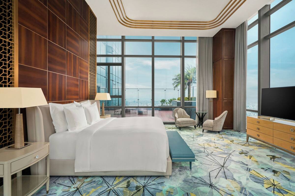 Una habitación de hotel moderna con una cama grande, dos mesitas de noche, un televisor y un balcón con vistas a la ciudad.
