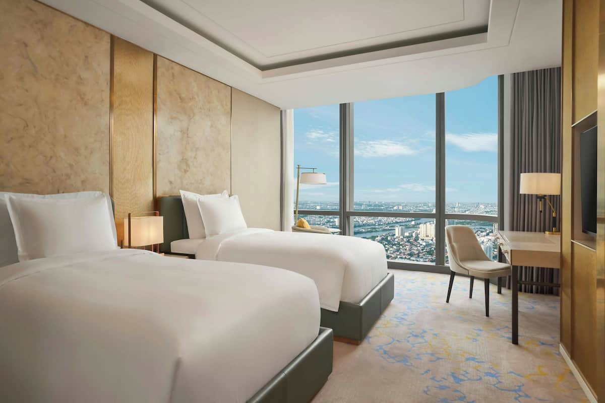 Habitación de hotel con dos camas, un escritorio, una silla y un ventanal con vistas a la ciudad.