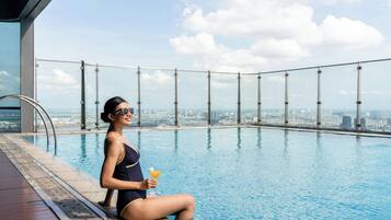 Una persona sentada junto a una piscina con vista a la ciudad, sosteniendo una bebida.