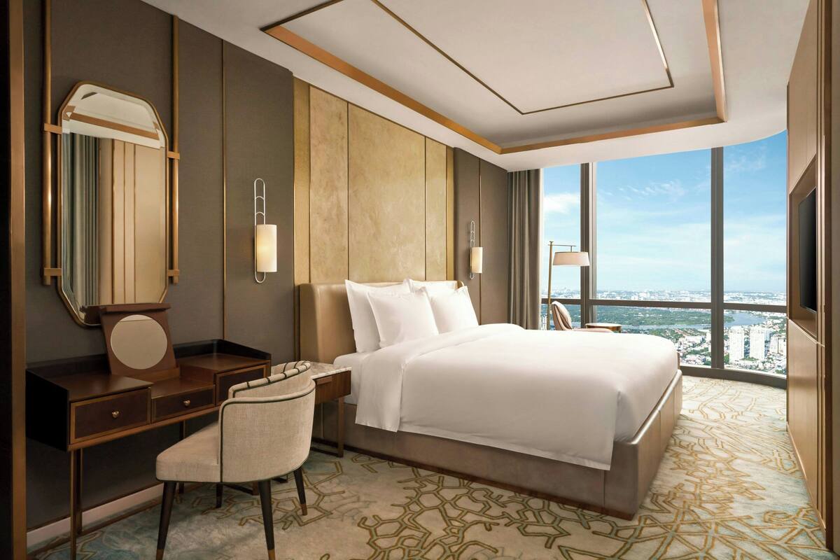 Una habitación de hotel moderna con una cama grande, un escritorio, una silla, un espejo y vistas de la ciudad a través de amplias ventanas.