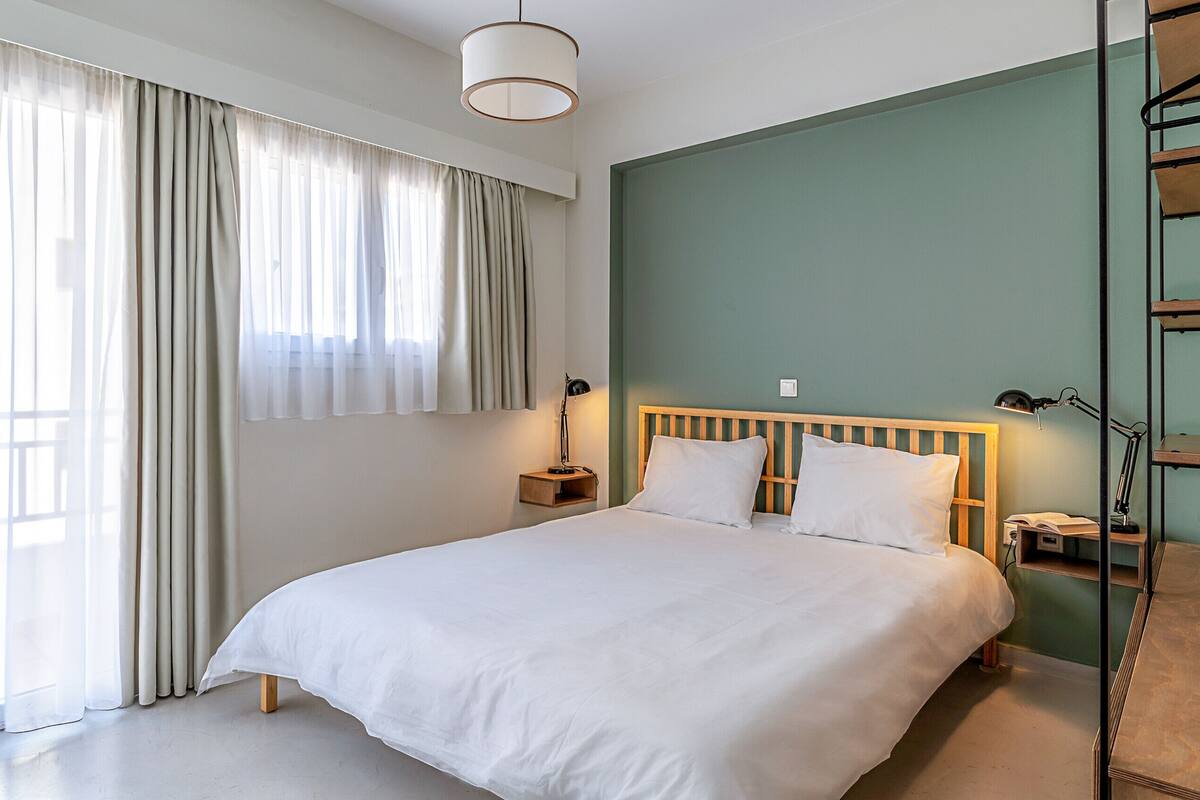A bedroom with a large bed, a bedside table with a lamp, and a window with curtains.
