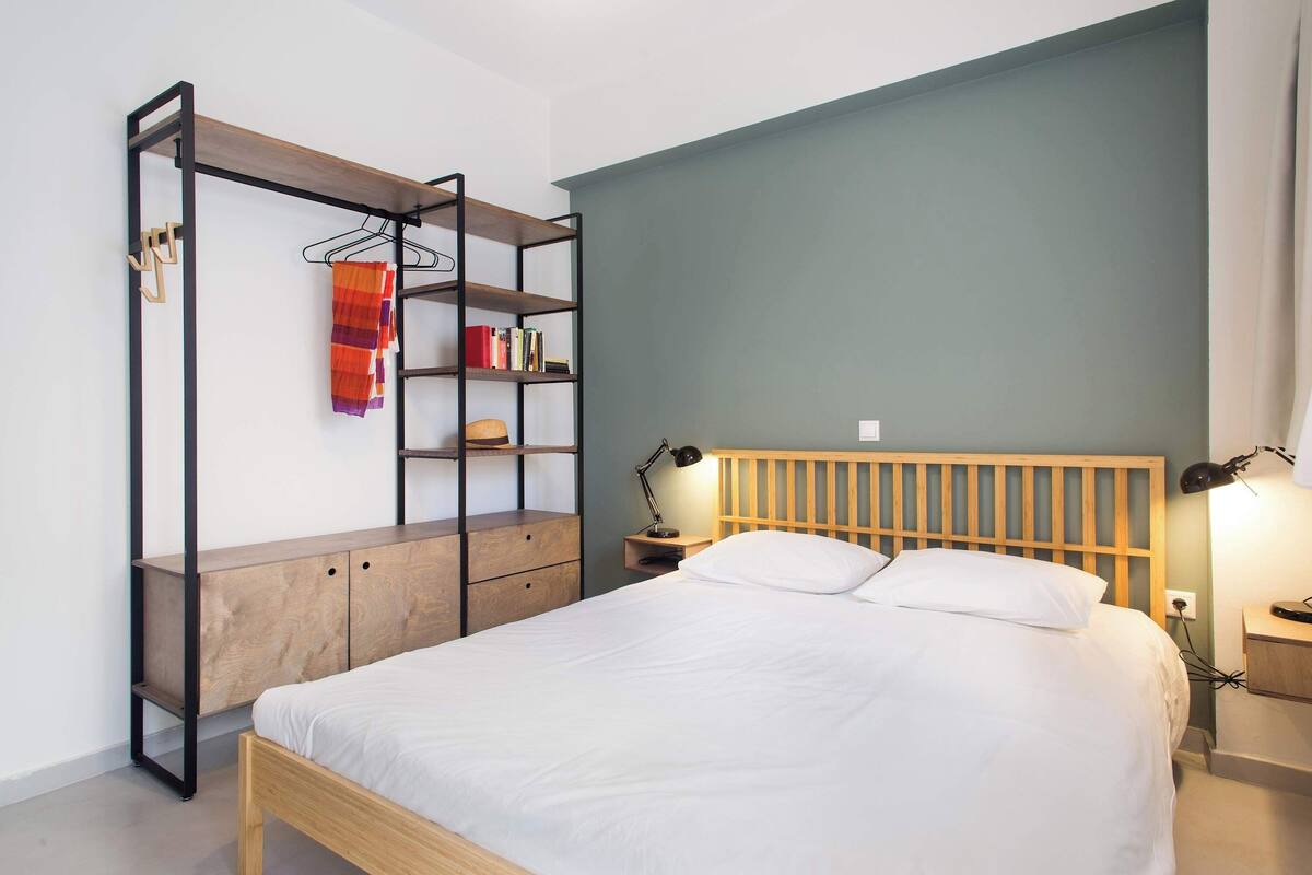 A bedroom with a bed, a wooden headboard, a nightstand, a shelf unit, and a hanging rack.