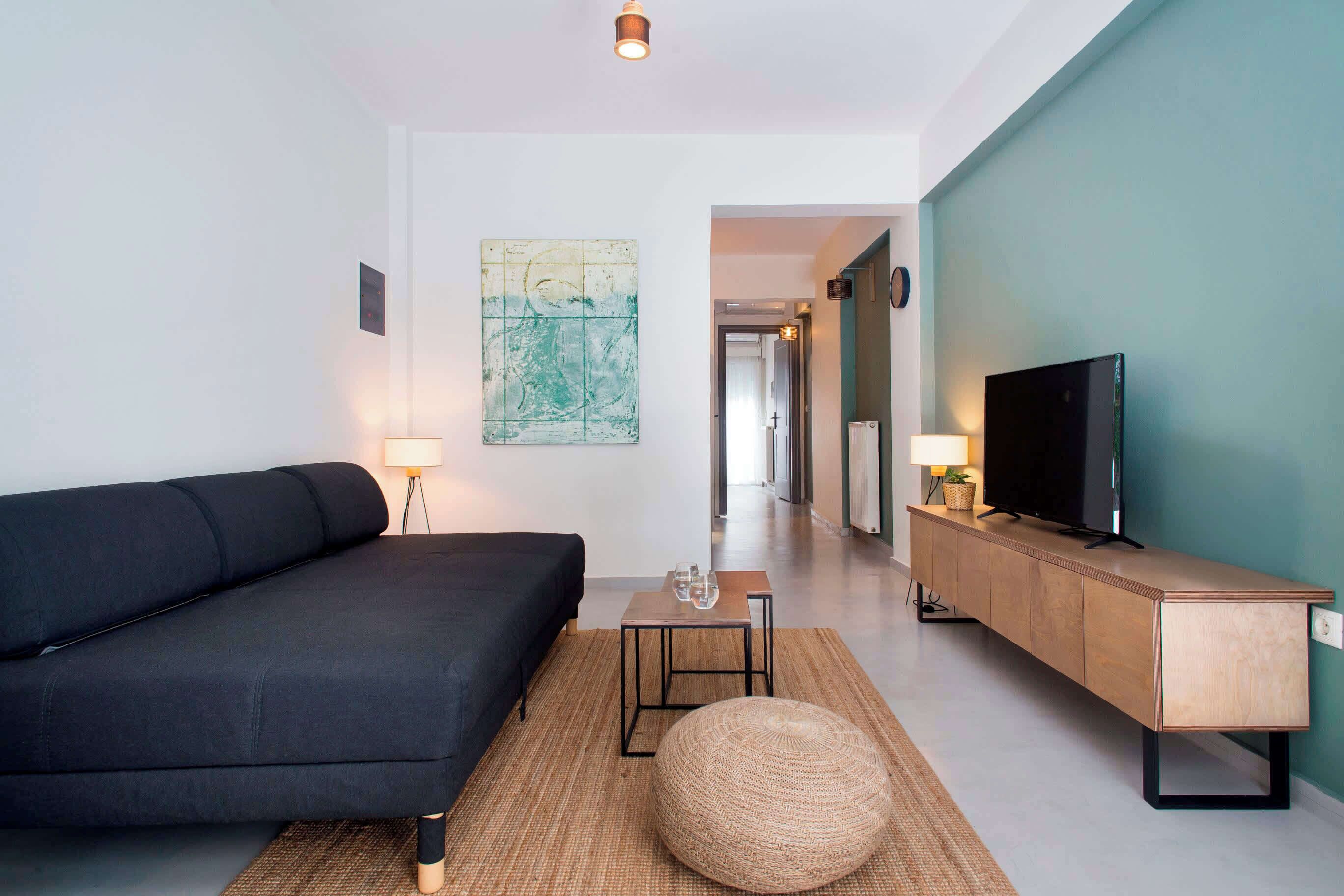 Una sala de estar moderna con un sofá negro, un mueble bajo para la televisión de madera y un otomán tejido.