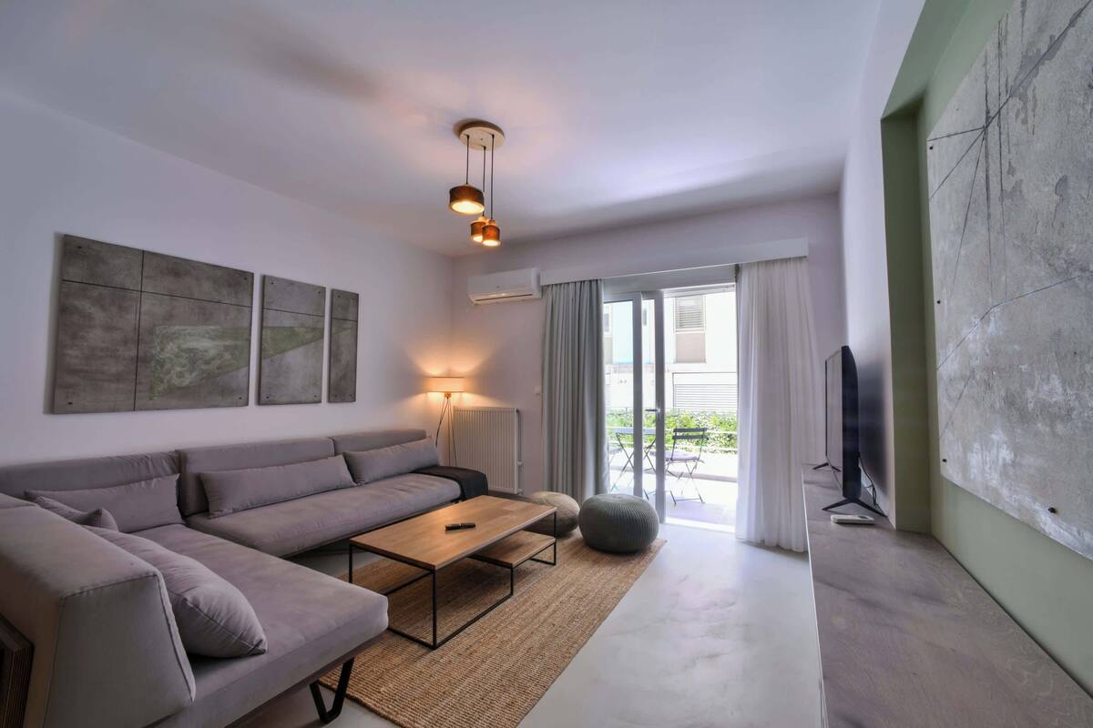 A modern living room with a sofa, coffee table, and a television. There is a large window with curtains and a view of greenery outside.