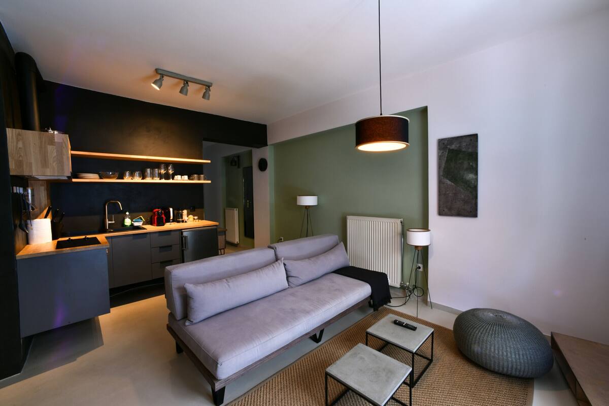 A modern living room with a grey sofa, a small coffee table, and a bean bag. A kitchen with a black countertop and shelves is visible in the background.