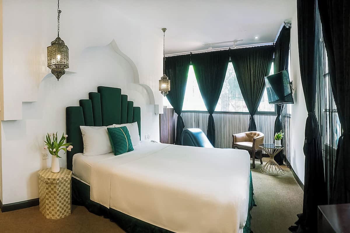 Une chambre d’hôtel avec un grand lit, une petite table de chevet ornée d’une plante, un élément décoratif mural et une grande fenêtre avec des rideaux.
