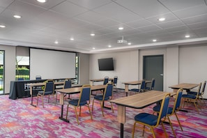A conference room with multiple tables and chairs, a projector screen, and a large window. - Home2 Suites by Hilton Portland Hillsboro (Hillsboro)
