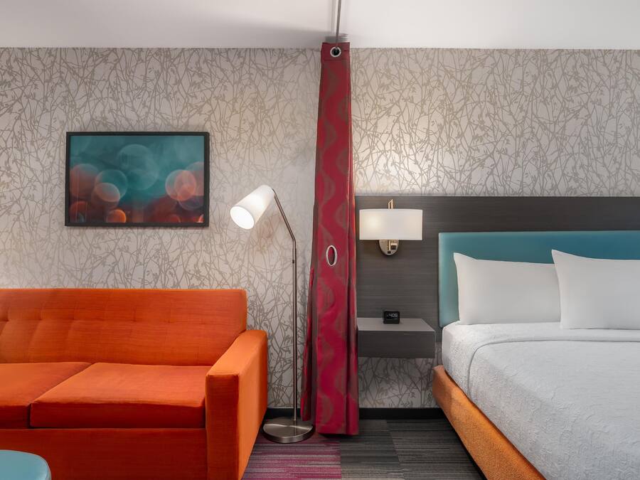 Una habitación de hotel con cama, mesita de noche, lámpara, silla y un cuadro en la pared.