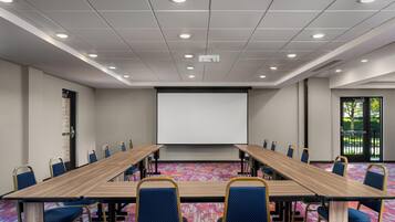 Un salón de conferencias con una larga mesa, sillas, una pantalla de proyector y una alfombra estampada.