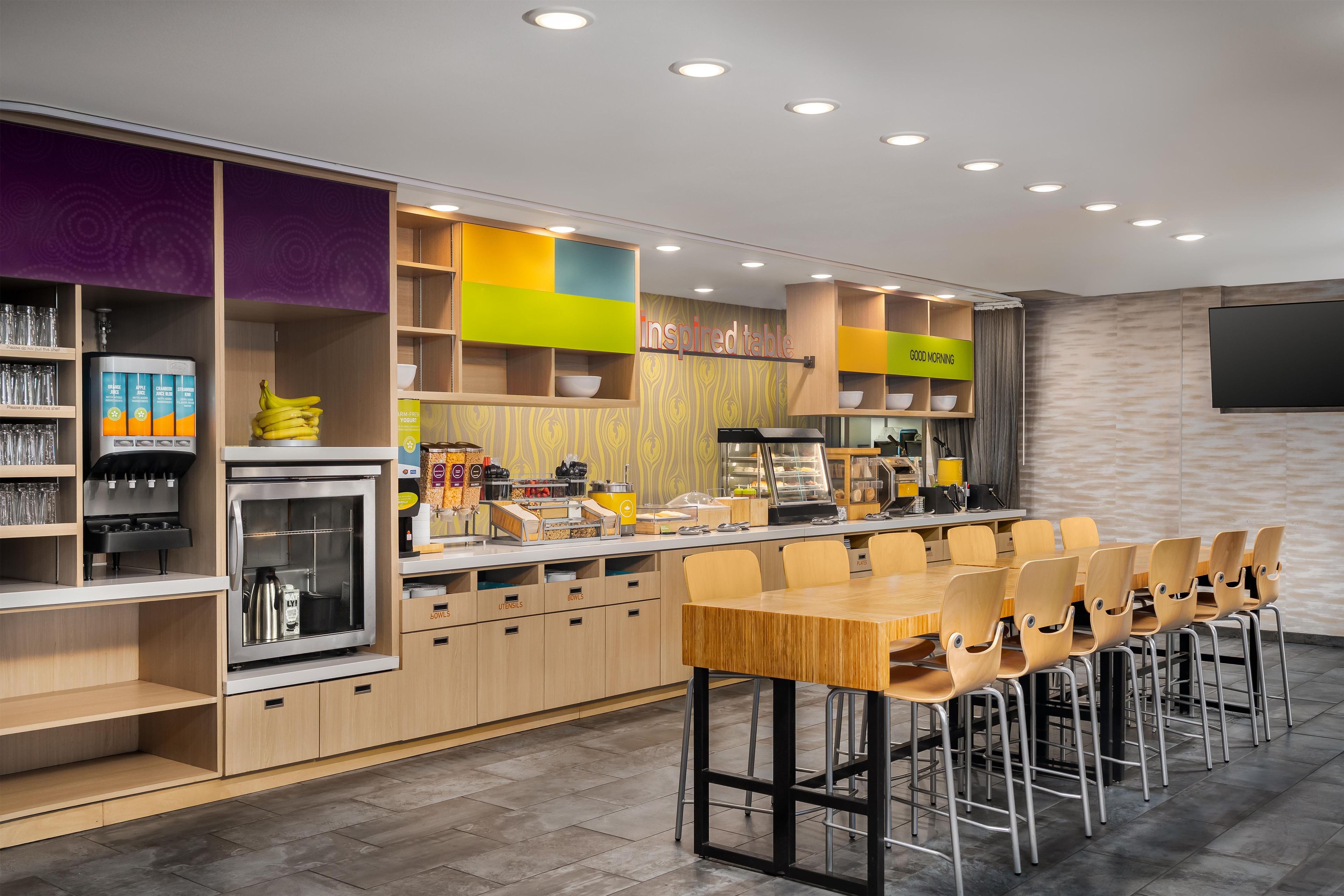 A modern café with a long wooden table and chairs, a coffee machine, and a variety of food items displayed.