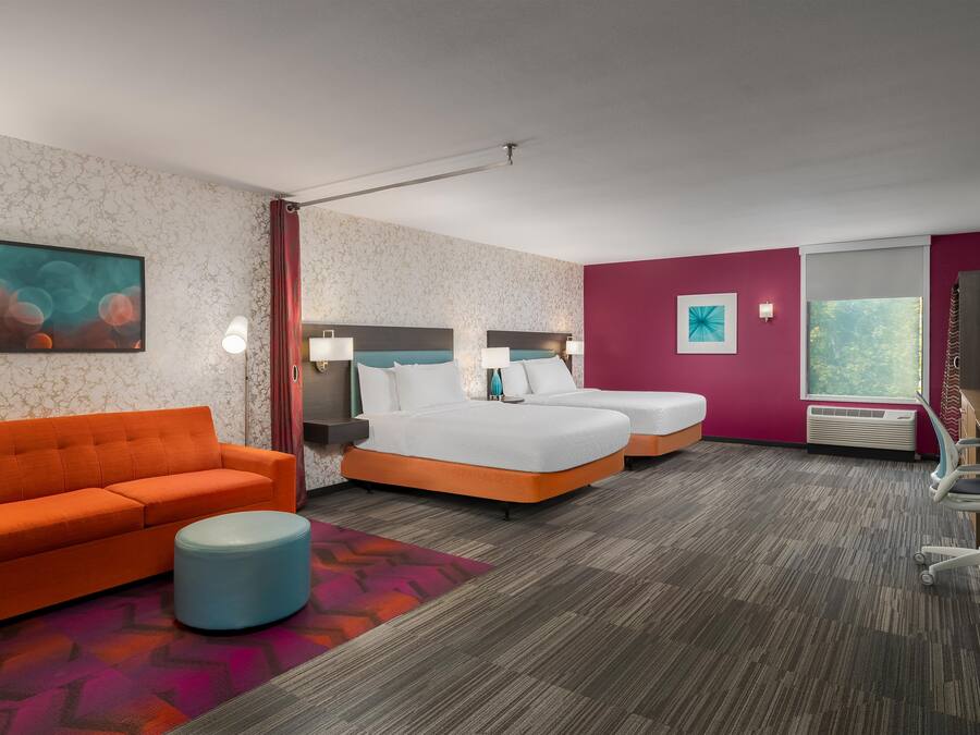 Quarto de hotel com sofá, duas camas, uma escrivaninha e uma janela.