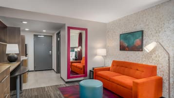 Quarto de hotel com sofá, um banquinho redondo, escrivaninha com luminária, espelho e uma pintura na parede.