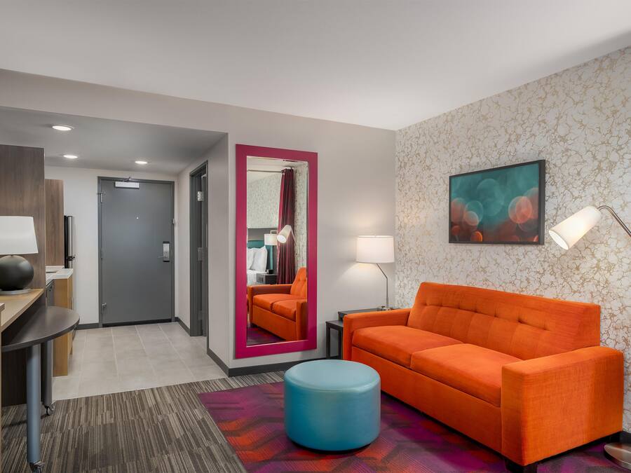 Quarto de hotel com sofá, um banquinho redondo, escrivaninha com luminária, espelho e uma pintura na parede.