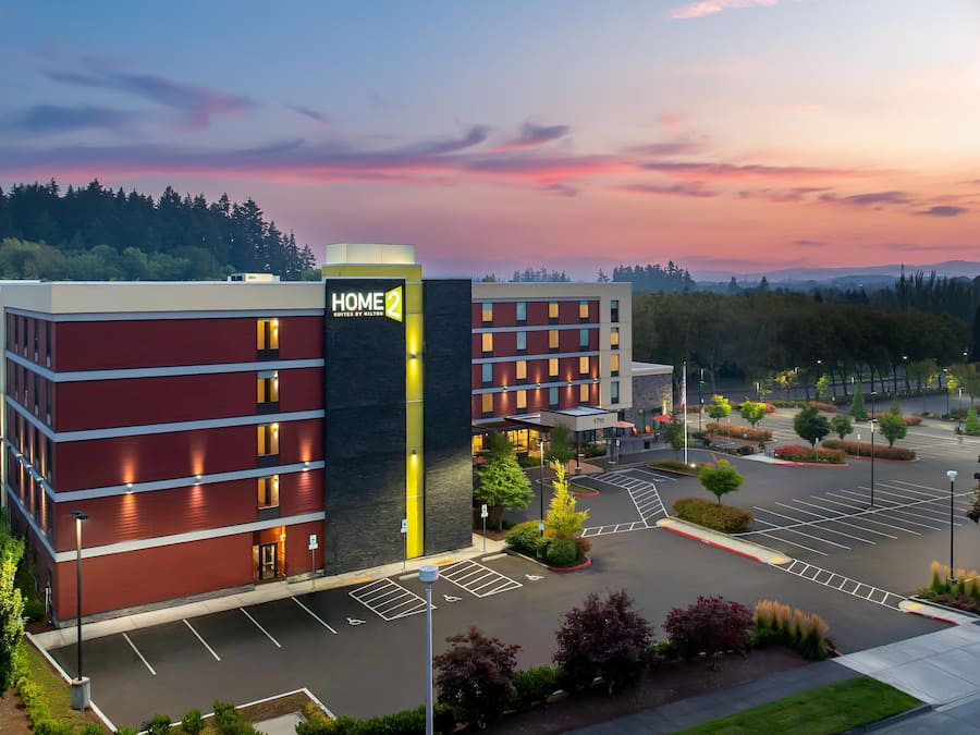 A hotel with a modern design, featuring a prominent 'HOME 2' sign, a parking lot, and a landscaped area with trees and shrubs.