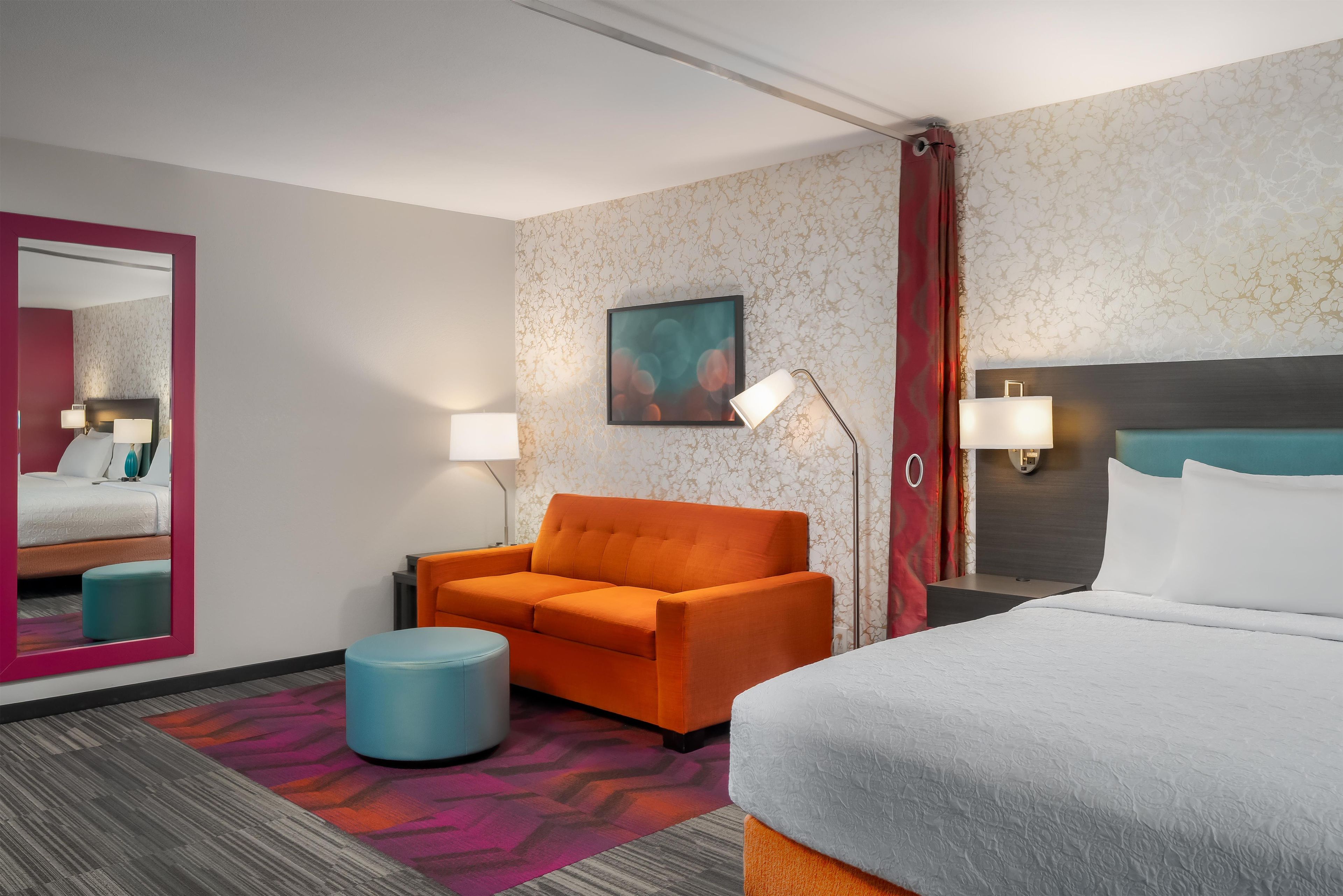 Una habitación de hotel con una cama, un sillón naranja, un reposapiés azul, una mesita de noche con una lámpara y un espejo grande.
