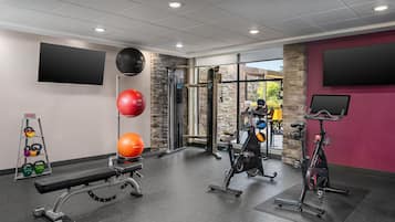 Academia com equipamentos de ginástica, um banco, kettlebells e uma TV fixada na parede.